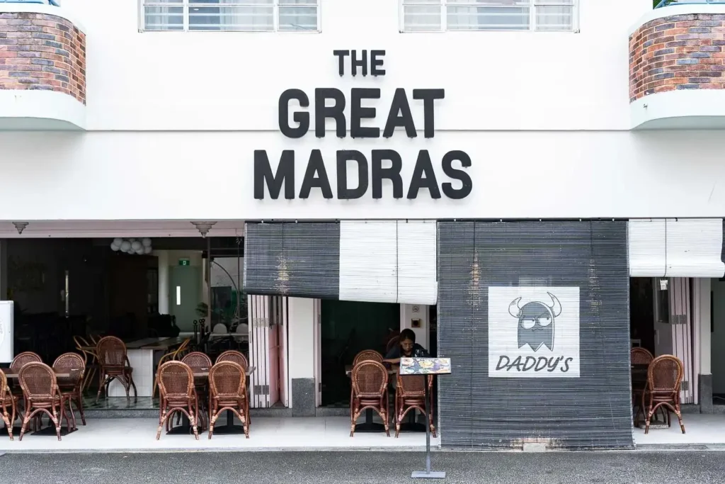 The exterior of The Great Madras hotel features bold black lettering on a clean white facade with Art Deco-inspired architecture. Below the signage, a bistro area is furnished with woven bistro chairs and tables tucked behind dark bamboo blinds.