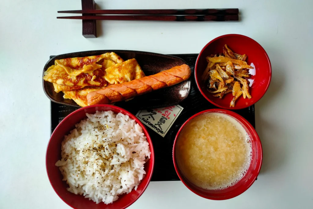 A top-down view shows a Japanese-style breakfast set served on a black tray with a pair of chopsticks resting at the top. The meal includes a large bowl of white rice seasoned with furikake, a bowl of miso soup, a side of fried small fish, and a plate with a rolled omelet and a scored sausage.