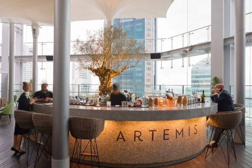 The image features an elegant rooftop bar named Artemis, which is highlighted by a warm, glowing counter and a central decorative tree adorned with lights. Set against a backdrop of city skyscrapers, the open-air space offers a sophisticated atmosphere with modern seating and large glass railings.