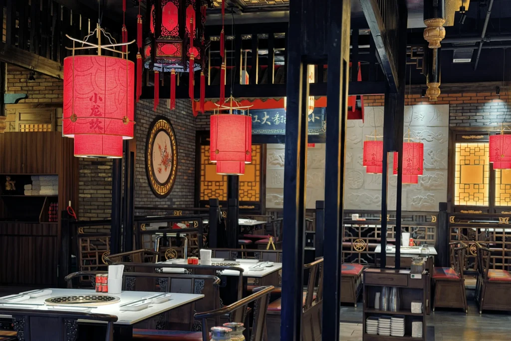 This restaurant features traditional Chinese decor, highlighted by vibrant red lanterns and dark wooden architectural elements. The interior is organized with white tables set for hot pot dining, surrounded by intricate brickwork and decorative wall panels.
