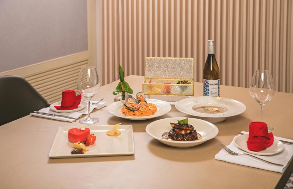 A sophisticated multi-course meal is elegantly arranged on a light wooden table, featuring dishes like seafood risotto, steak, and a heart-shaped dessert. The setting is complete with a bottle of Chardonnay, two wine glasses, and vibrant red napkins folded into rose shapes.