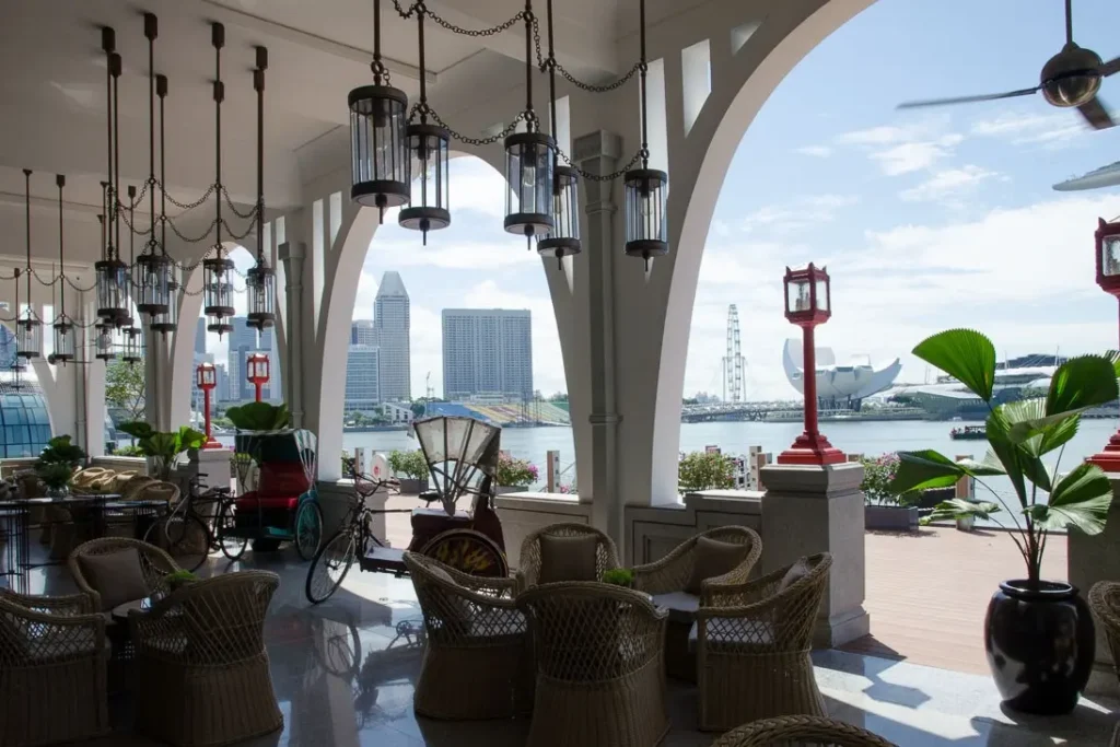 An elegant, open-air restaurant terrace features hanging lantern chandeliers, wicker seating, and traditional trishaws parked near the columns. Through the large arched openings, a scenic waterfront view reveals the Singapore skyline, including the ArtScience Museum and the Singapore Flyer.