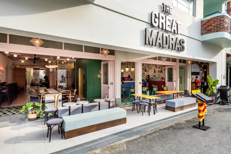 The Great Madras is a vibrant hotel with a stylish outdoor seating area featuring long benches and colorful tables under a modern sign. Its open-air design blends retro aesthetic elements with contemporary urban architecture.