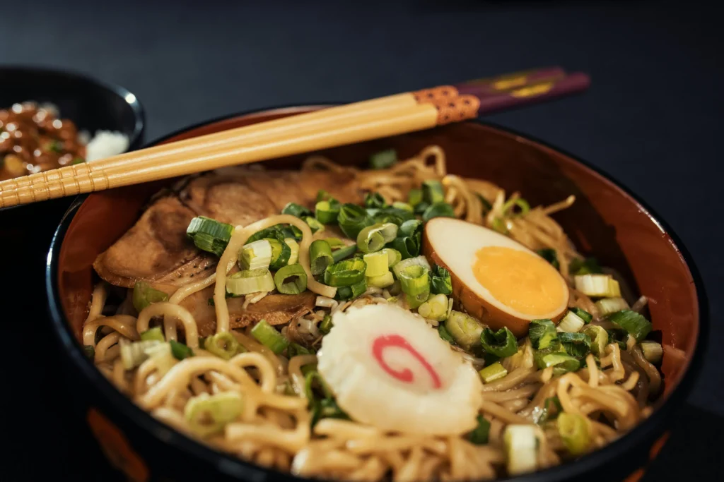 A close-up shot features a steaming bowl of ramen topped with a swirl-patterned narutomaki fish cake, a soft-boiled soy egg, chashu pork, and chopped green onions. Wooden chopsticks rest across the rim of the dark bowl against a muted, dark background.