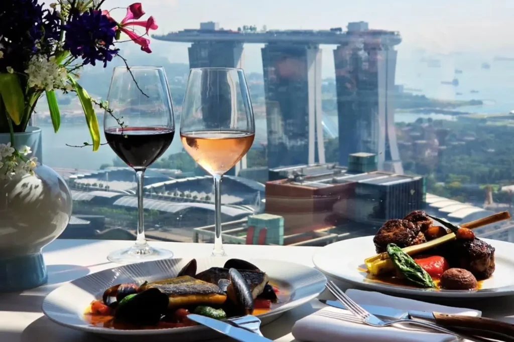 The image shows a sophisticated dining table set with elegant seafood and meat dishes accompanied by two glasses of wine. In the background, a large window offers a stunning panoramic view of the Marina Bay Sands hotel and the surrounding cityscape in Singapore.