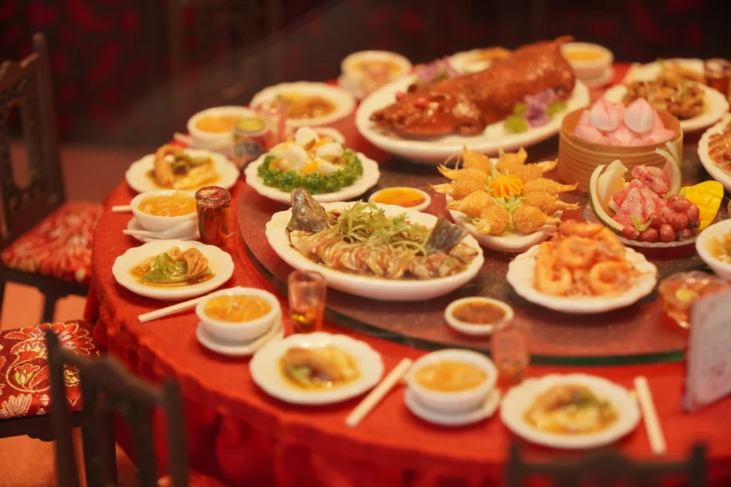 A large round table draped in red cloth is set for a lavish Chinese banquet, featuring miniature versions of dishes like roasted suckling pig, steamed fish, and dim sum. The scene captures an intricate traditional feast, complete with small chairs, tiny chopsticks, and a rotating glass lazy susan at the center.