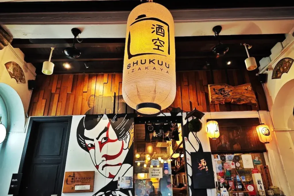 An outdoor shot displays the entrance of Shukuu Izakaya, featuring a large, glowing paper lantern with the restaurant's name hanging above the doorway. The facade is adorned with a striking, large mural of a traditional Japanese Kabuki actor alongside wooden paneling and smaller decorative lanterns.
