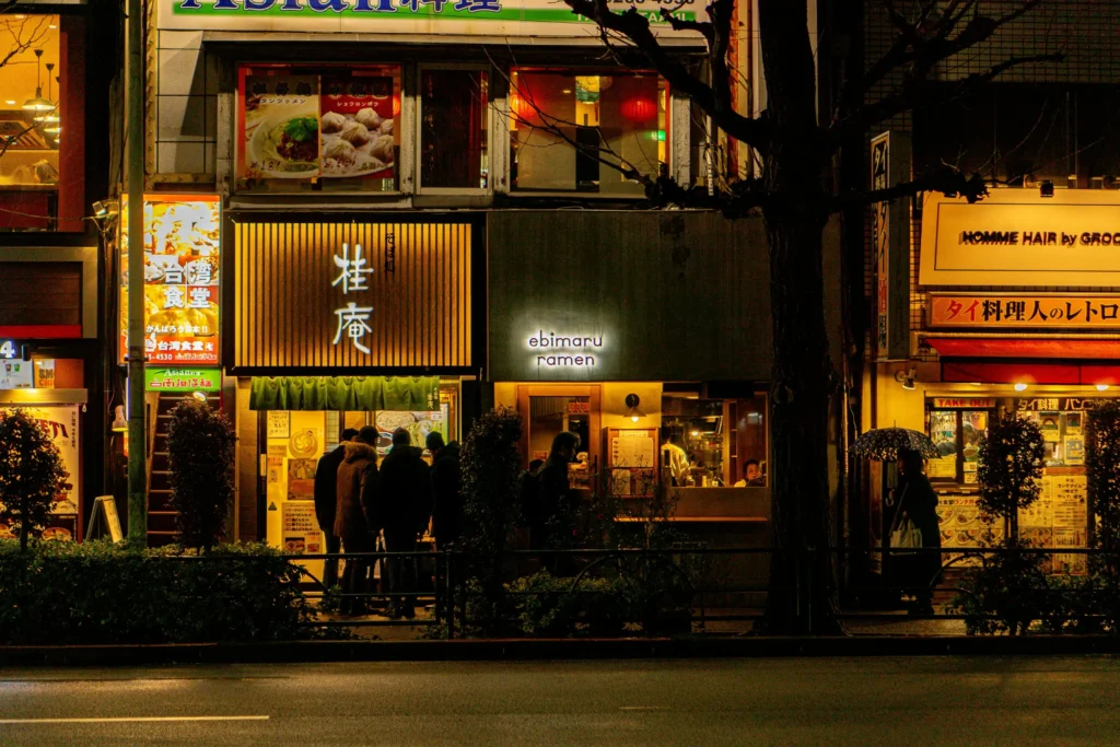 A night scene captures a lively Tokyo street lined with illuminated restaurant storefronts, including a brightly lit noodle shop with Japanese kanji and an adjacent eatery labeled "ebimaru ramen." A few people stand outside the entrances under the warm glow of the signs, while a large tree and low green bushes frame the sidewalk in the foreground.