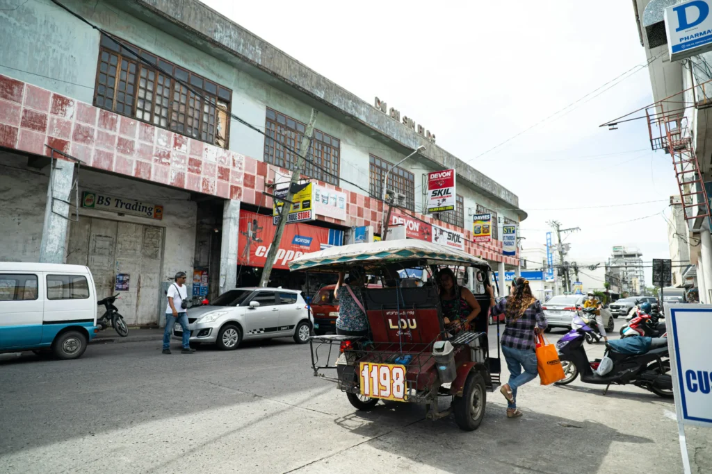 The image shows a bustling street scene in front of a weathered, multi-story commercial building with various storefront signs. In the foreground, a motorized tricycle taxi carries passengers past parked cars and scooters on a bright, sunny day.