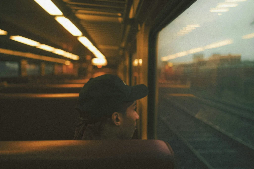 A side-profile view of a person wearing a dark baseball cap sitting inside a dimly lit train car and looking out the window. The interior is bathed in warm, grainy light from overhead fixtures, while train tracks and a hazy outdoor landscape are visible through the glass.