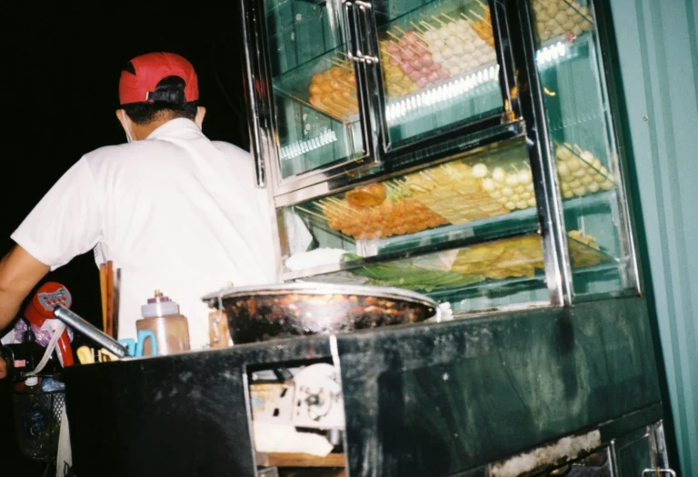 A street food vendor in a red cap and white shirt stands beside a glass display case filled with various colorful skewers. The brightly lit cart, positioned outdoors at night, features organized rows of fish balls and street snacks ready for cooking.