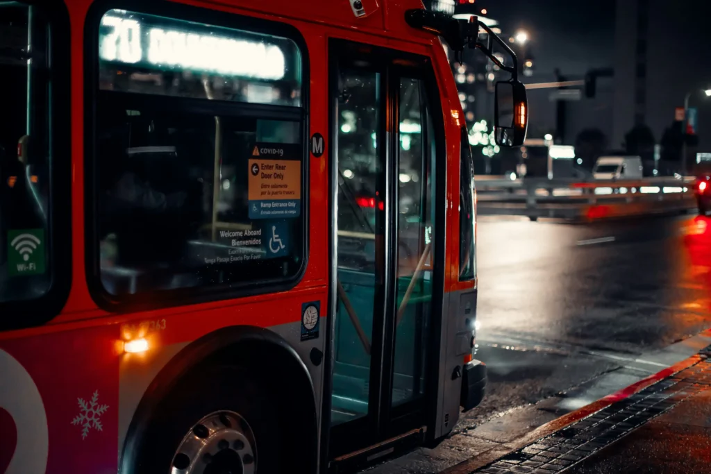 A vibrant red and white bus is parked along a wet city street at night, its interior lights casting a warm glow through the windows. The scene captures a moody, urban atmosphere with blurred city lights and reflections shimmering on the damp pavement.