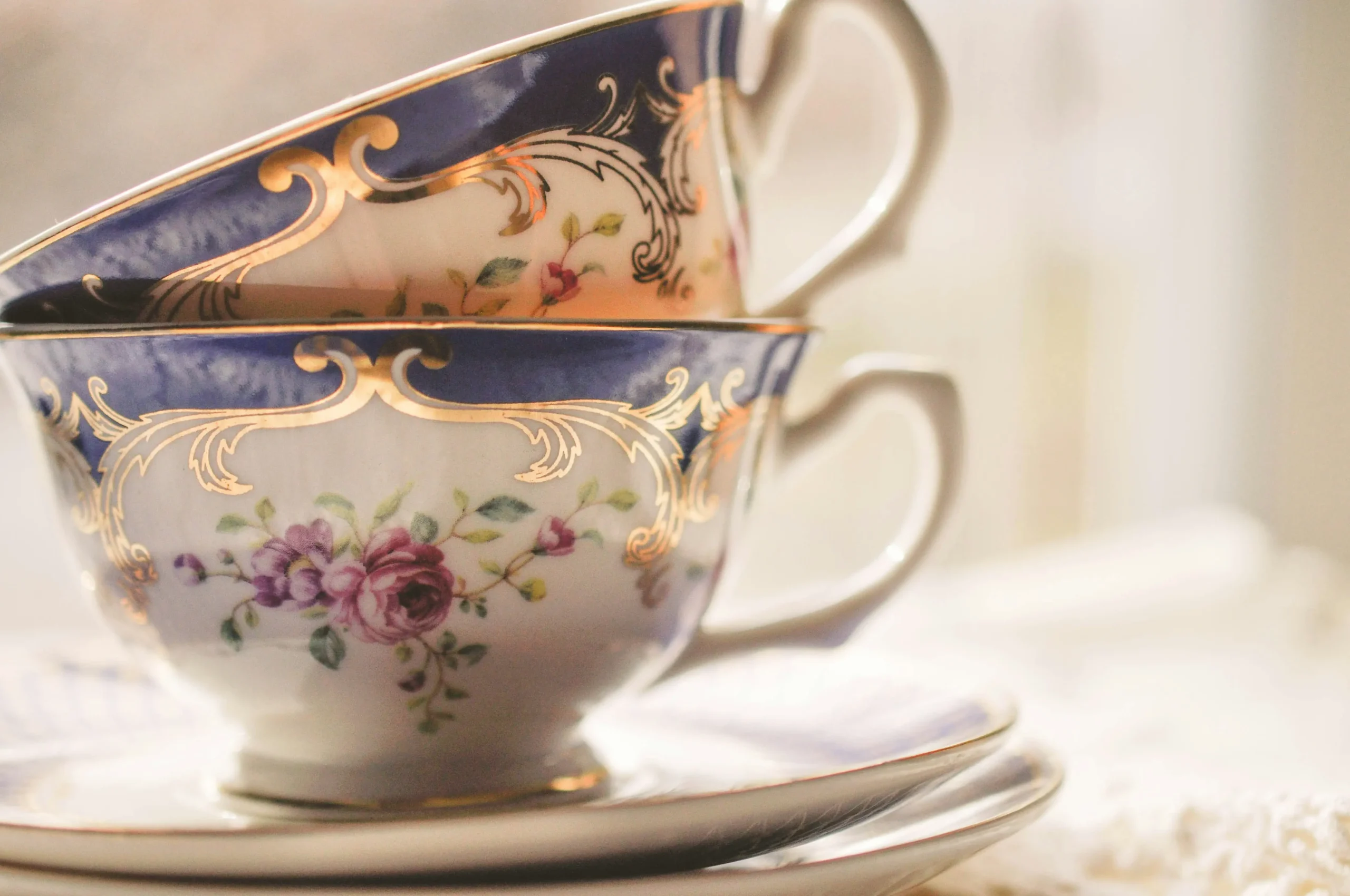 Two vintage porcelain teacups with intricate gold detailing and floral patterns are stacked on top of matching saucers. The cups feature a deep blue rim with elegant gold filigree and delicate pink and purple rose illustrations on a white background.