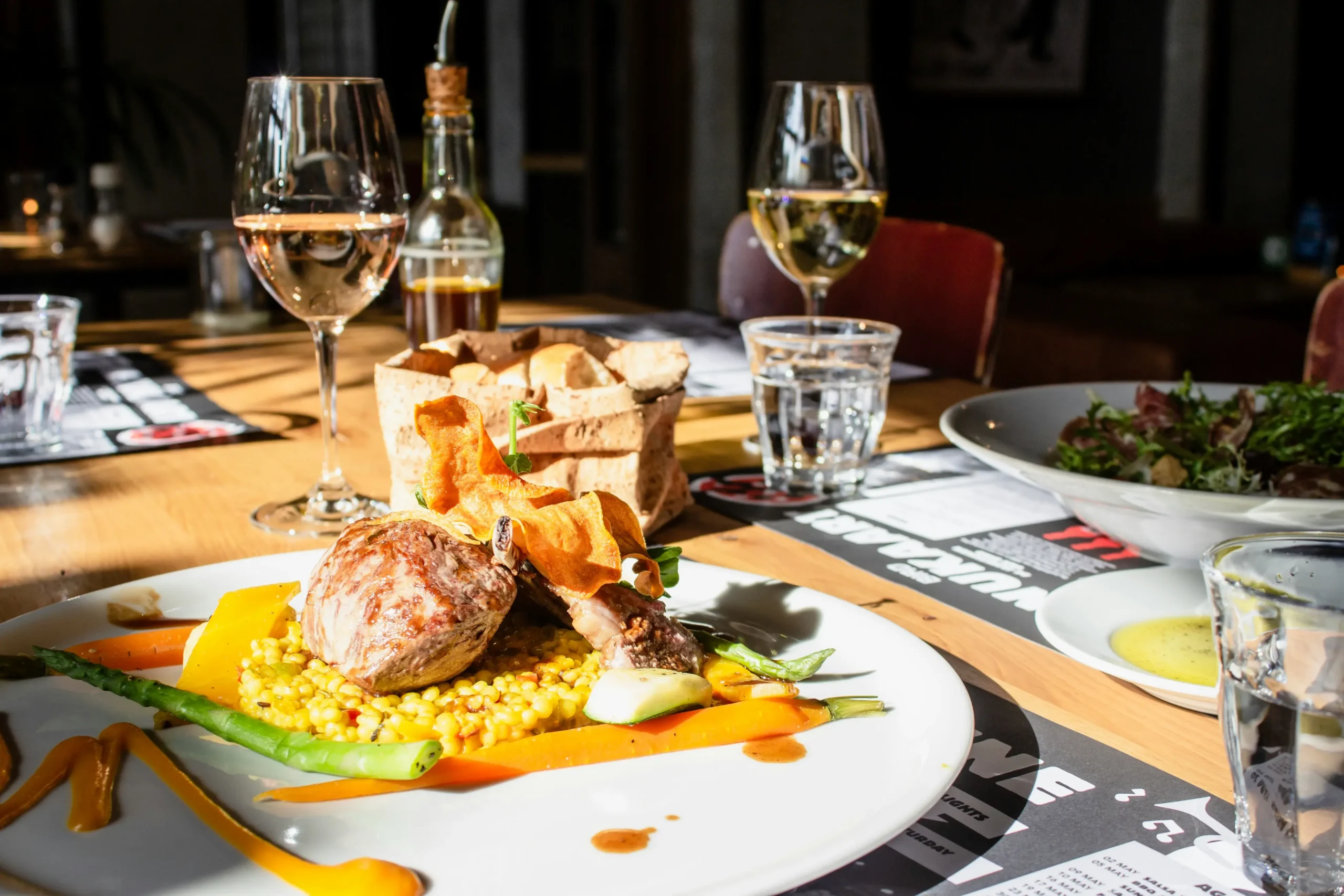 A beautifully plated gourmet meal featuring a roasted cut of meat, vibrant vegetables, and yellow grains sits on a sunlit restaurant table. In the background, wine glasses, bread, and a side salad complete the upscale dining setting.
