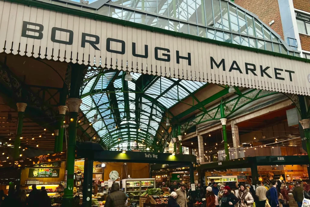 A large, white wooden sign reads "BOROUGH MARKET" in bold black lettering across the entrance of a bustling indoor market. Beneath the distinctive green iron and glass roof, numerous people walk between illuminated food stalls such as "Ted's Veg" and "RAYA".