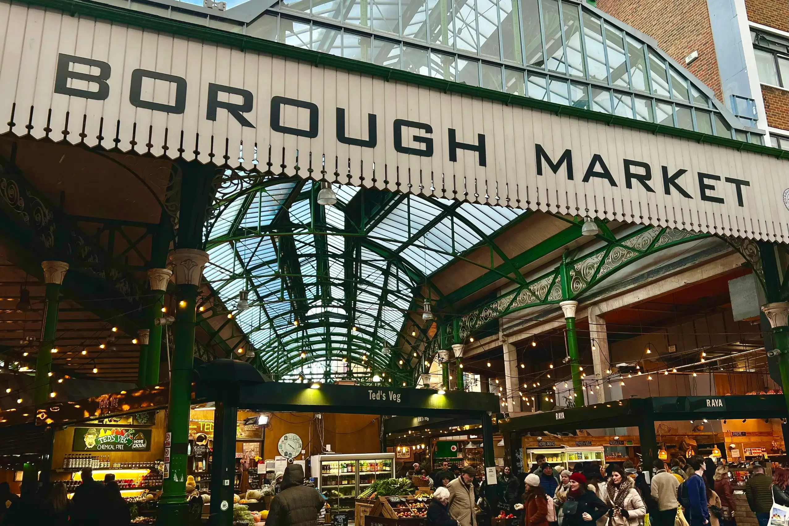 A large, white wooden sign reads "BOROUGH MARKET" in bold black lettering across the entrance of a bustling indoor market. Beneath the distinctive green iron and glass roof, numerous people walk between illuminated food stalls such as "Ted's Veg" and "RAYA".