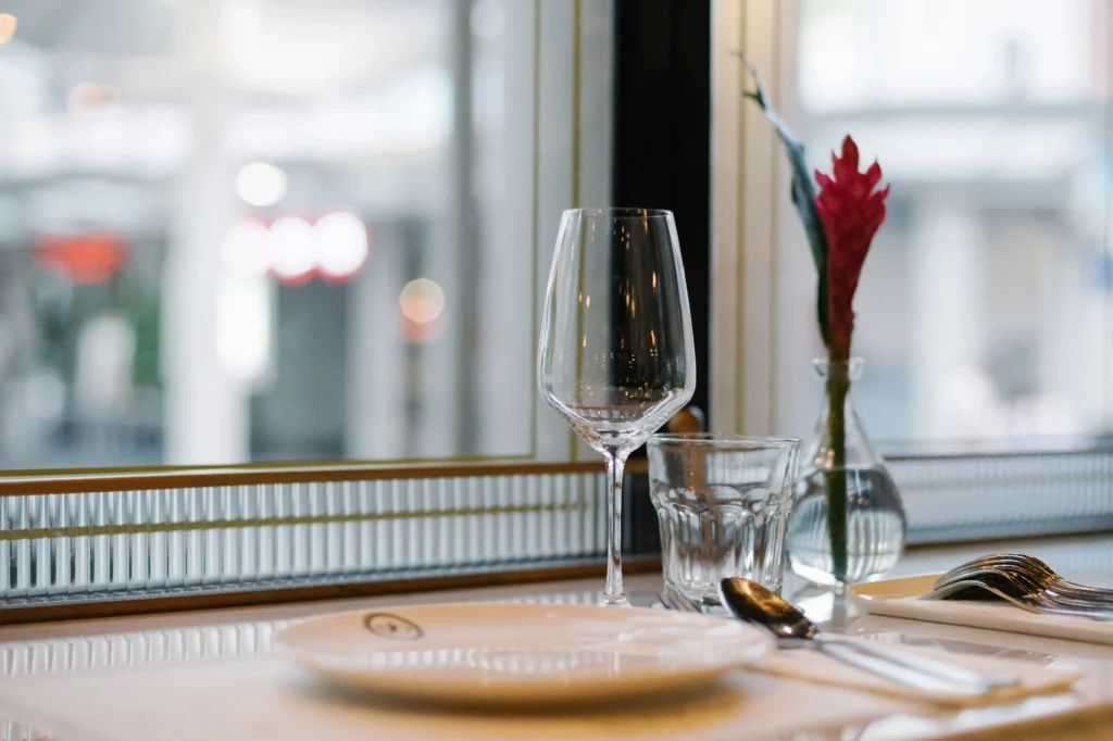 A close-up shot captures an elegant table setting next to a window, featuring a white plate, a wine glass, a water glass, and silverware. A small glass vase holding a single bright pink flower adds a touch of color against the softly blurred background of an outdoor street view.