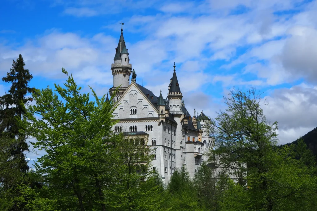 A fairytale-style white stone castle with tall, pointed spires rises above a dense canopy of lush green trees under a bright blue sky filled with scattered white clouds. The structure features intricate architectural details, multi-tiered windows, and dark-tiled roofing nestled against a mountainous backdrop.