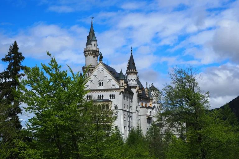 A fairytale-style white stone castle with tall, pointed spires rises above a dense canopy of lush green trees under a bright blue sky filled with scattered white clouds. The structure features intricate architectural details, multi-tiered windows, and dark-tiled roofing nestled against a mountainous backdrop.