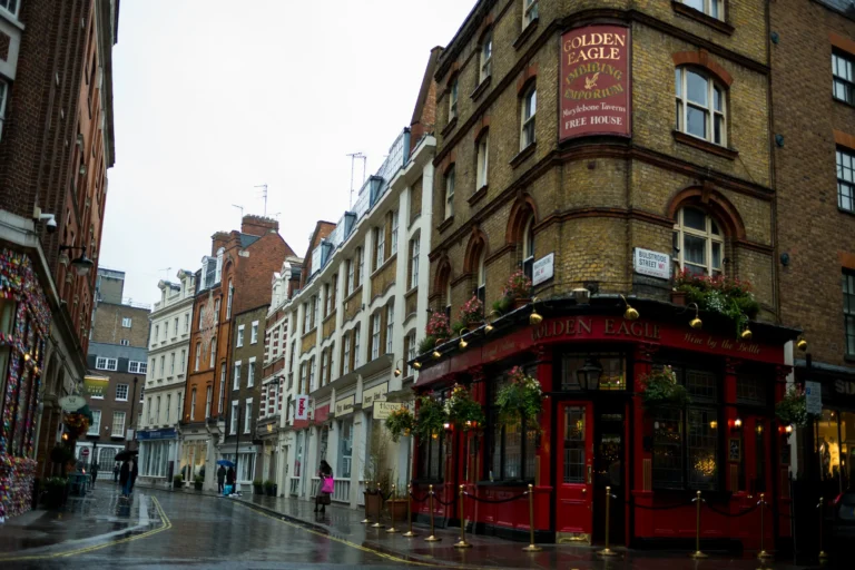 A wet, rainy street in London curves past a traditional multi-story brick pub named "The Golden Eagle" on Bulstrode Street. The pub features a vibrant red storefront, hanging flower baskets, and a large sign identifying it as an "Imbibing Emporium" and "Free House."