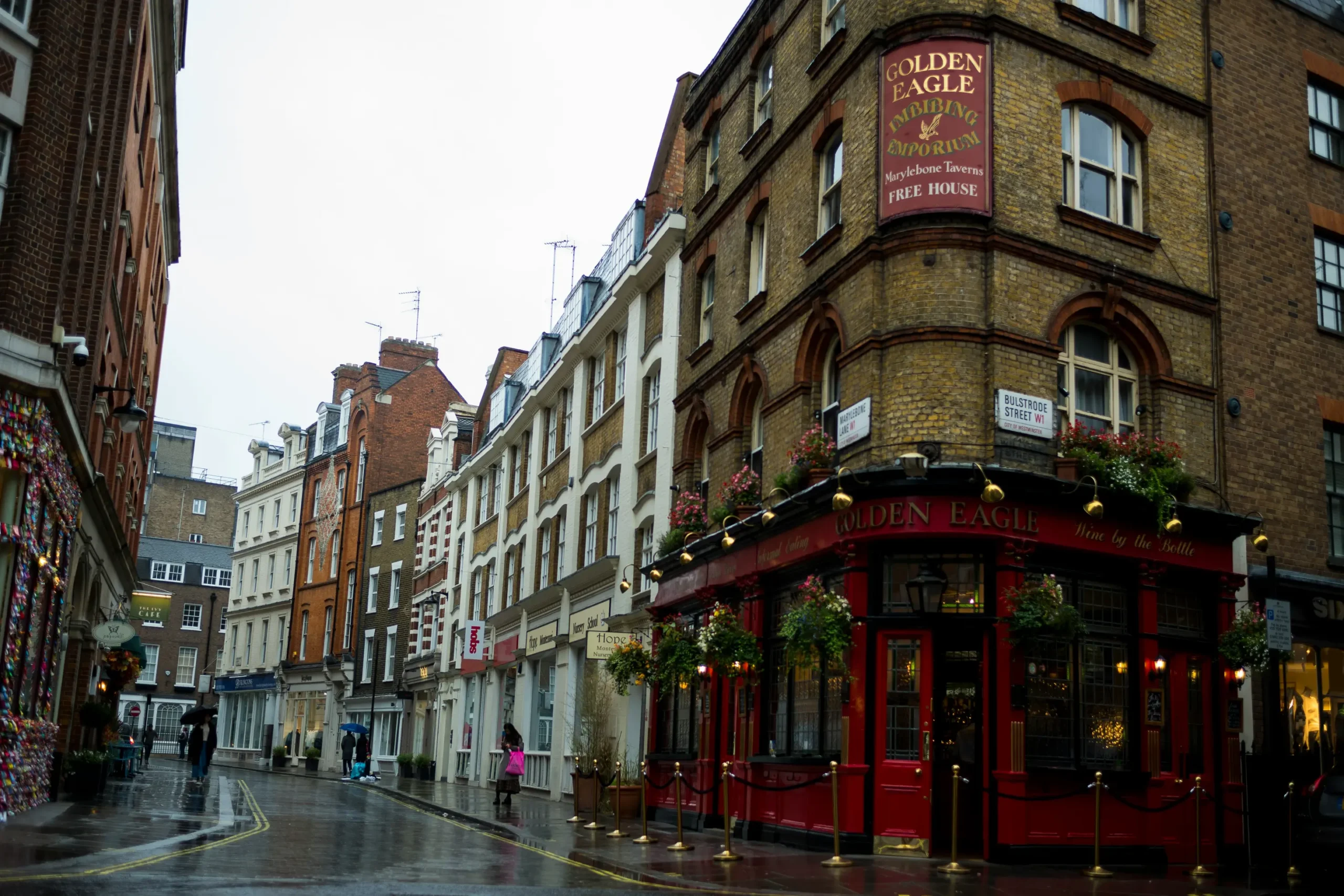 A wet, rainy street in London curves past a traditional multi-story brick pub named "The Golden Eagle" on Bulstrode Street. The pub features a vibrant red storefront, hanging flower baskets, and a large sign identifying it as an "Imbibing Emporium" and "Free House."