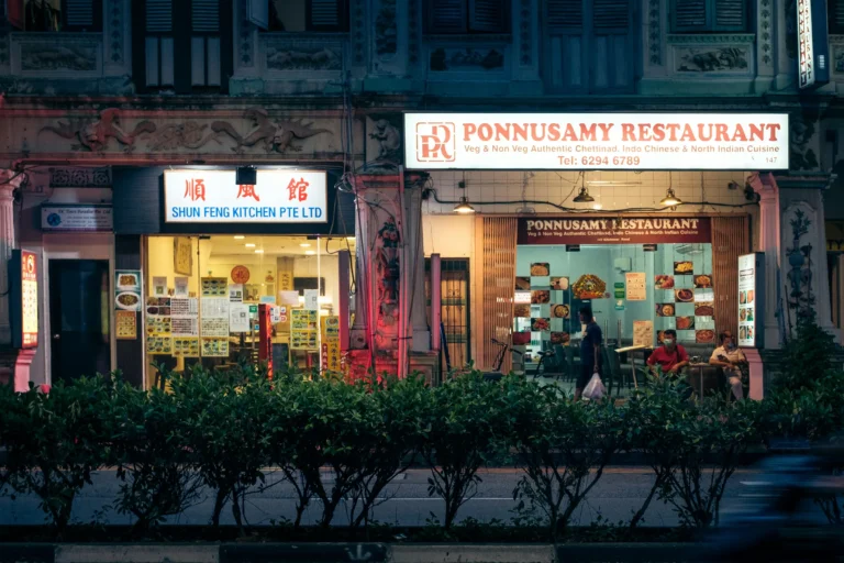 A nighttime street view shows the brightly lit storefronts of Shun Feng Kitchen Pte Ltd, a Chinese restaurant, and Ponnusamy Restaurant, an Indian eatery, side by side in a traditional building. A low row of green hedges lines the foreground in front of the restaurants, where a few people can be seen inside and near the entrances.