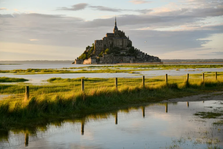 A distant view of Mont Saint-Michel, a historic tidal island abbey in Normandy, France, rising above the surrounding water under a soft, overcast sky. In the foreground, a grassy green field with a wooden fence and reflective puddles catches the warm glow of the setting sun.