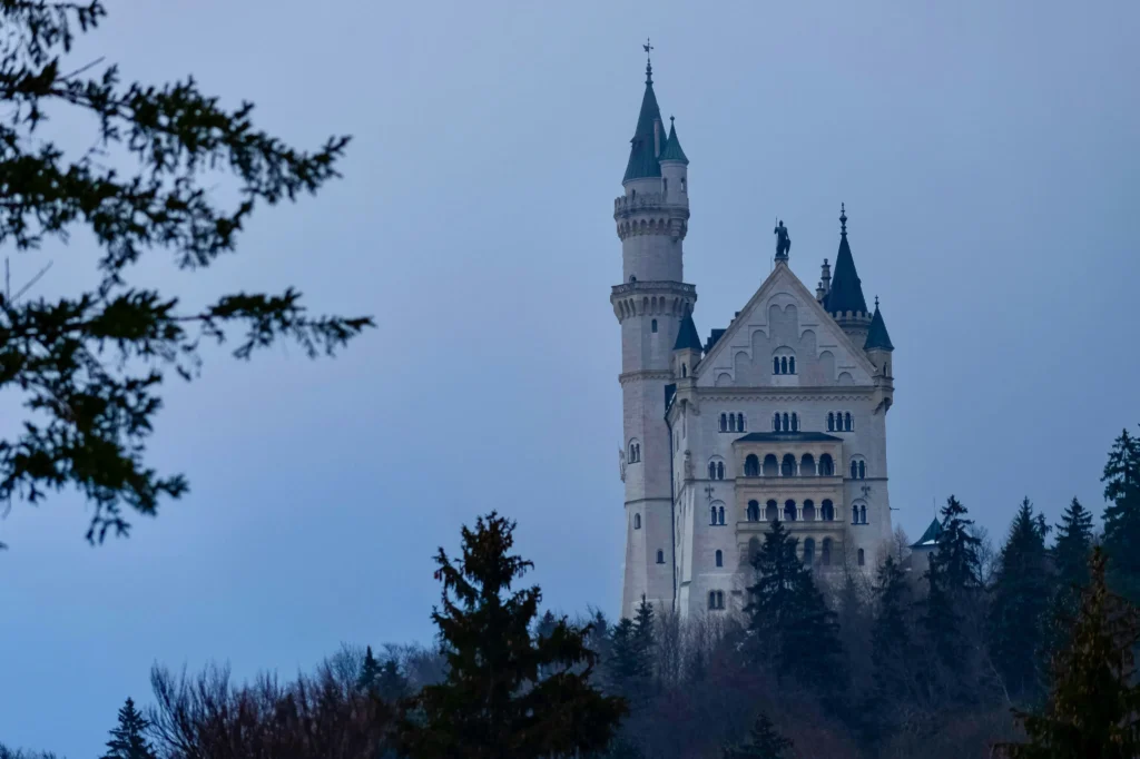 A majestic white stone castle with tall, pointed green spires stands prominently on a forested hill under a hazy blue sky. Dark evergreen trees frame the bottom and left sides of the shot, emphasizing the castle's isolated, fairy-tale appearance.