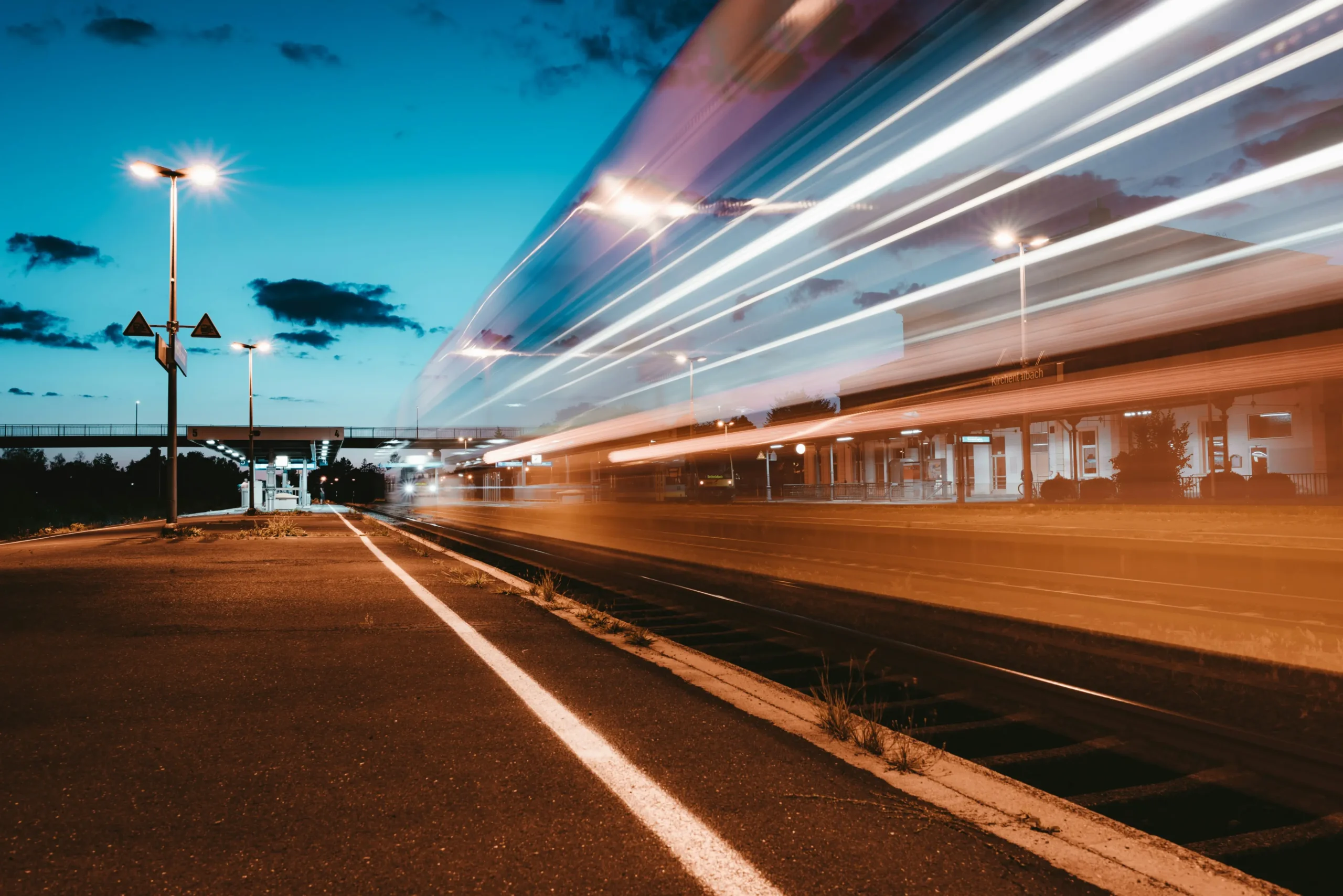 A long-exposure photograph captures a train speeding past an outdoor station at dusk, leaving behind bright streaks of white and orange light. The empty, illuminated platform in the foreground contrasts sharply with the deep blue, cloudy twilight sky above.