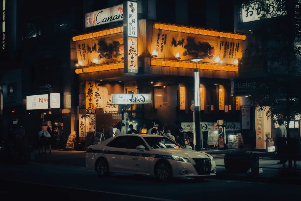 A white taxi sits parked on a dark street at night in front of a multi-story Japanese building illuminated by warm, glowing orange lanterns and signage. Pedestrians gather near the brightly lit storefronts and entrances under the vibrant city lights.