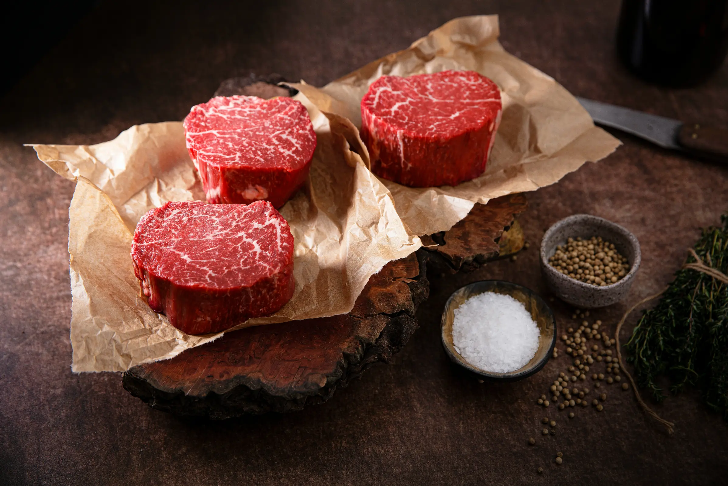 Three thick, raw filet mignon steaks with fine white marbling sit on pieces of brown butcher paper over a rustic wooden board. Accompanying the meat are small bowls of coarse sea salt, whole white peppercorns, a bundle of fresh thyme, and a knife handle in the background.