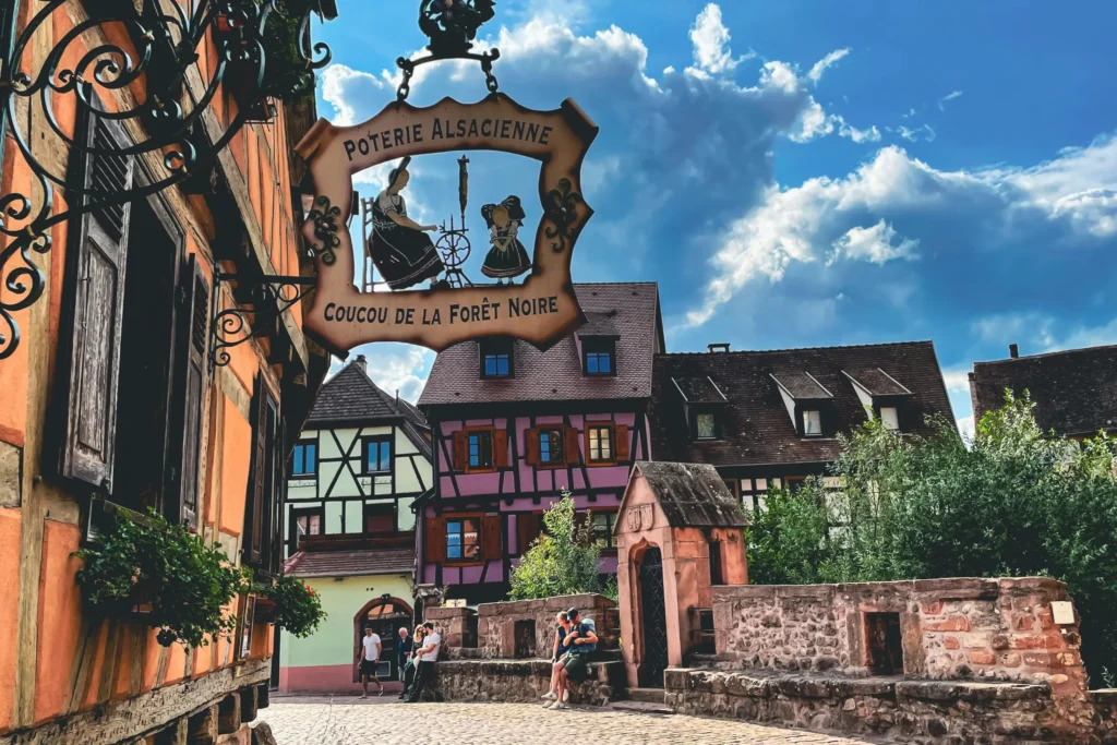 A low-angle view shows an ornate wrought-iron sign for an Alsatian pottery and Black Forest cuckoo clock shop hanging above a narrow, cobbled street lined with traditional half-timbered houses in various colors. In the background, people relax near a stone bridge under a bright, partly cloudy sky.