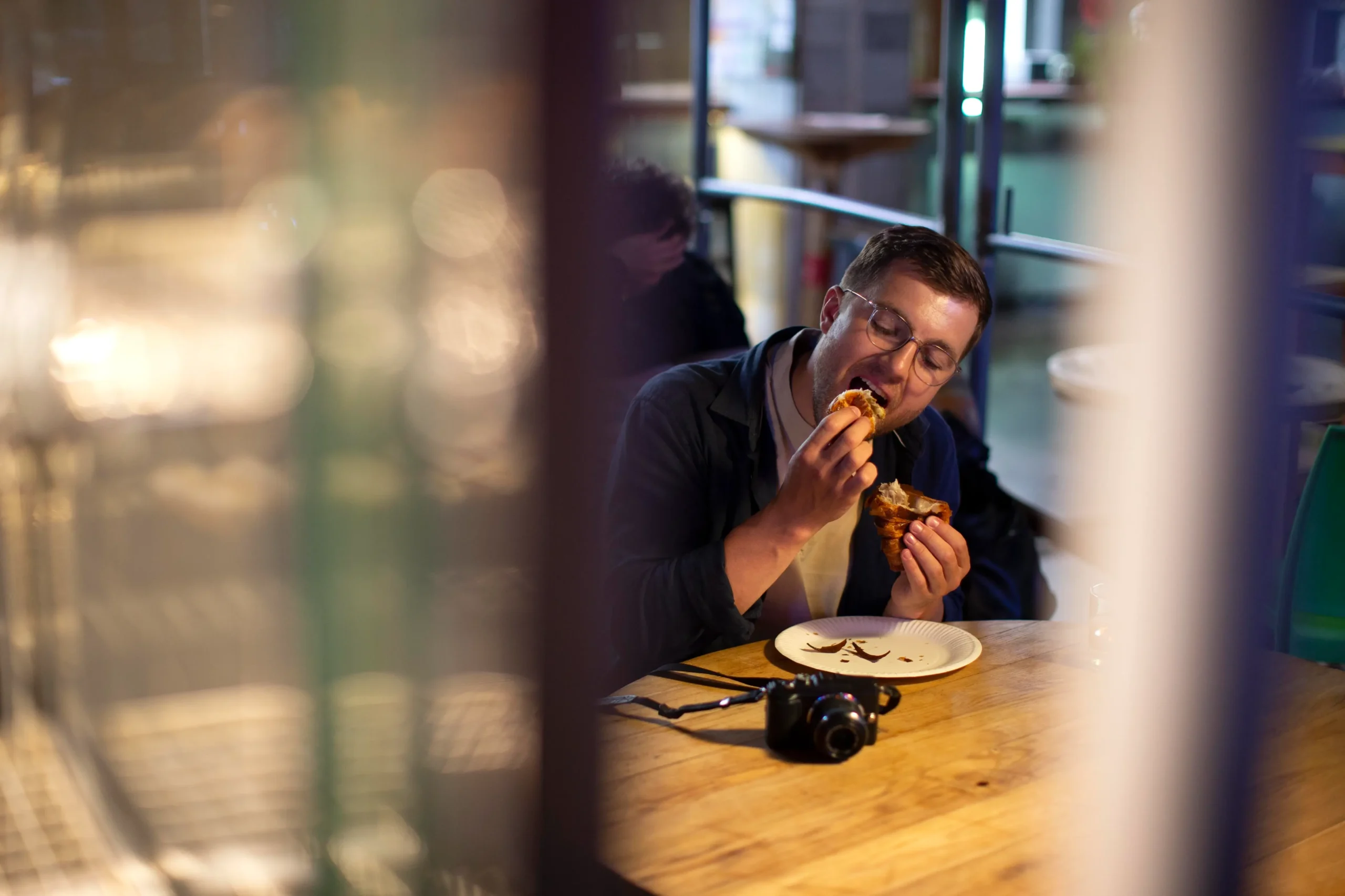 Candid street photography of a man with glasses eating a pastry at a round wooden table, viewed through a blurry glass window with warm bokeh reflections.