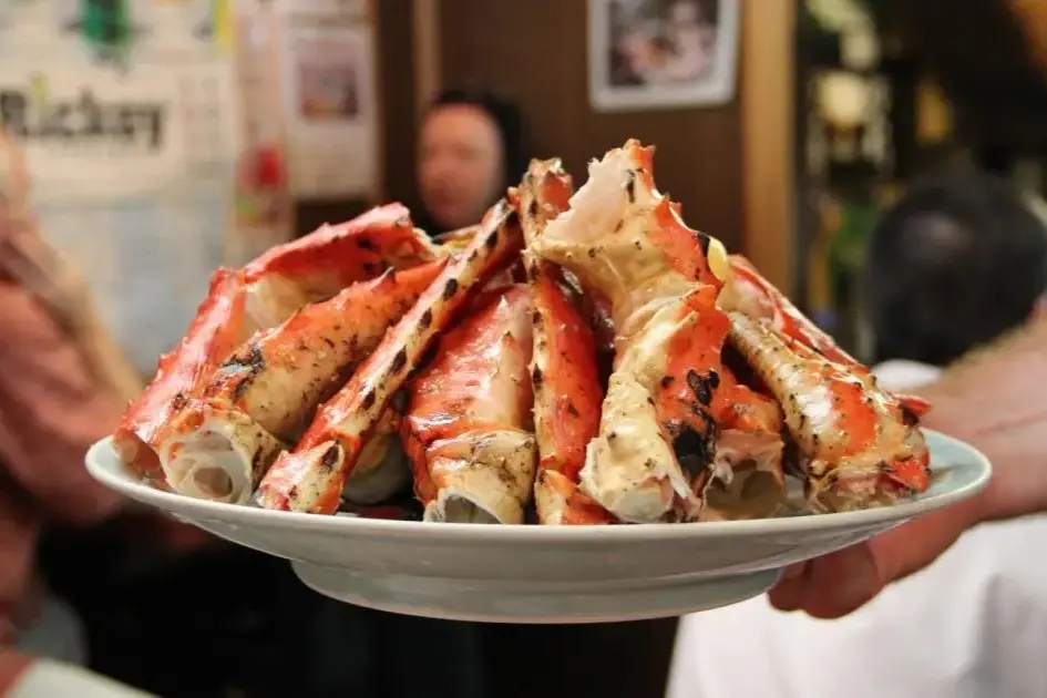 A large platter piled high with cooked, grilled king crab legs is being held up in a bustling restaurant setting. The image showcases the bright red and white shells, lightly charred from cooking, filling the center of the frame against a softly blurred background.