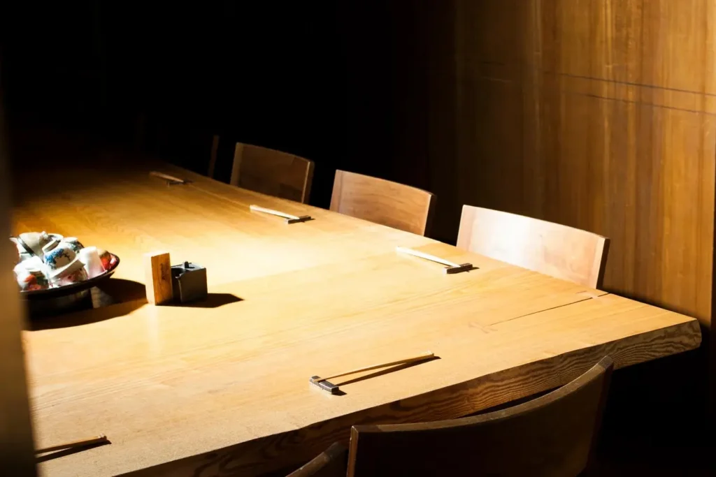 The image features a long, light-colored wooden dining table illuminated by a warm spotlight in a dimly lit room with wooden paneled walls. Wooden chairs line the table, which is set with pairs of chopsticks on rests and a bowl filled with ceramic cups on one end.