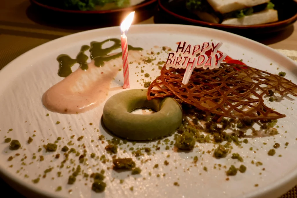 A beautifully plated matcha-flavored dessert donut sits on a white plate decorated with a lit birthday candle and a "Happy Birthday" topper. The plate is artistic, featuring green crumbs, a swipe of light pink sauce, and a delicate, net-like chocolate lattice.