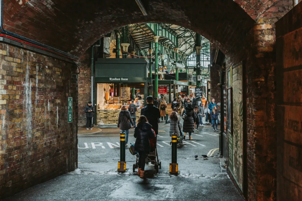 A view looking out from beneath a dark, weathered brick archway onto a bustling outdoor market filled with people. In the background, pedestrians walk near stall fronts under a large green metal canopy structure, including one stall labeled "Wyndham House."
