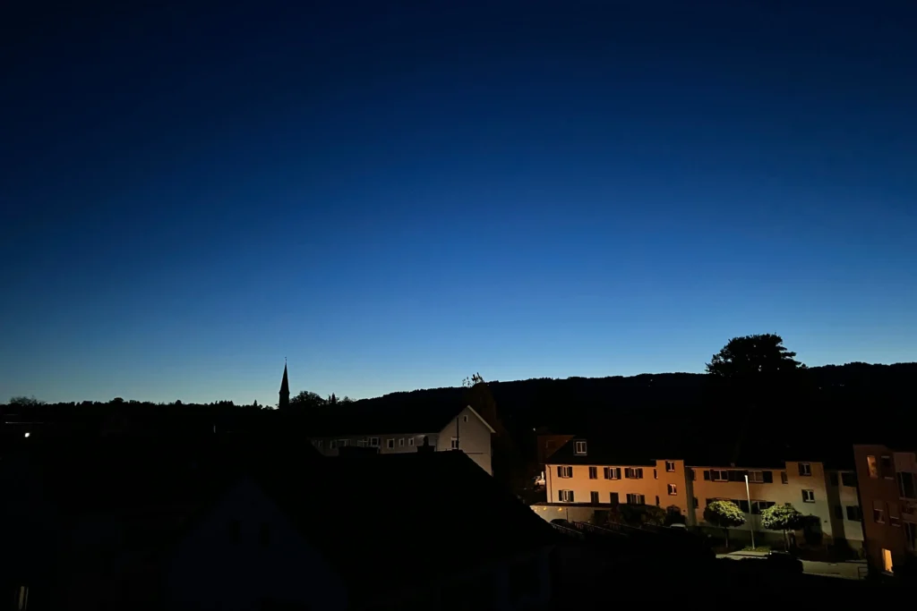 A serene night view of a small town silhouette under a deep blue gradient twilight sky. In the foreground, warm light glows from a few building windows, contrasting against the dark outlines of rooftops and a distant church steeple.