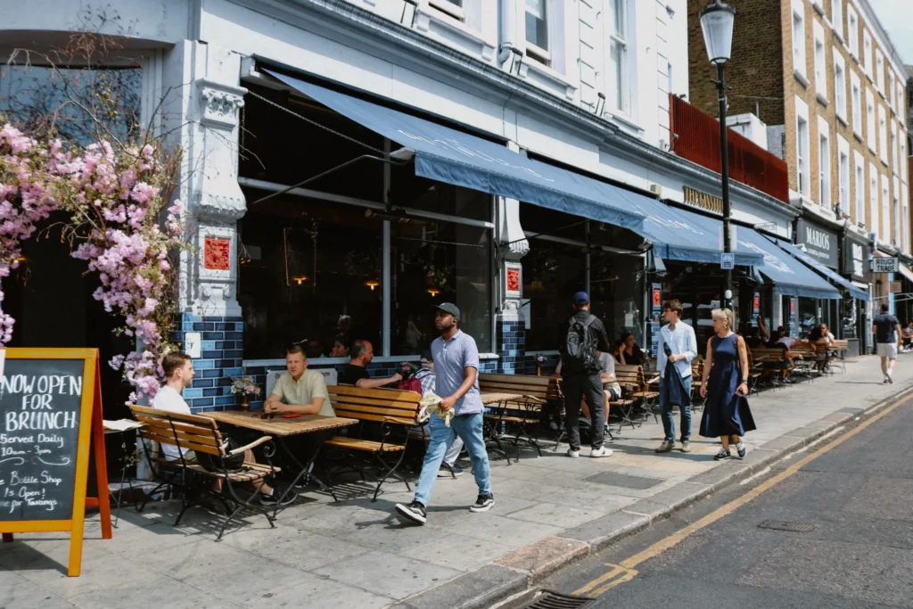 A bustling city sidewalk features patrons sitting at outdoor wooden tables underneath a large blue awning of a restaurant. Pedestrians stroll along the walkway on a sunny day next to a chalkboard sign advertising brunch.