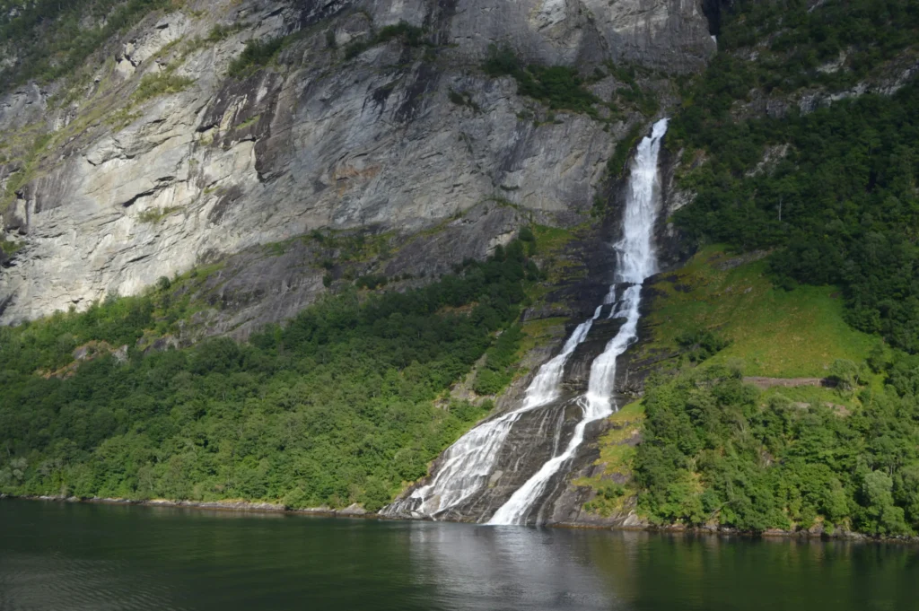 A large waterfall cascades down a steep, rocky cliffside and empties directly into a calm body of water below. Dense green trees and lush vegetation cover the lower slopes surrounding the rushing water.