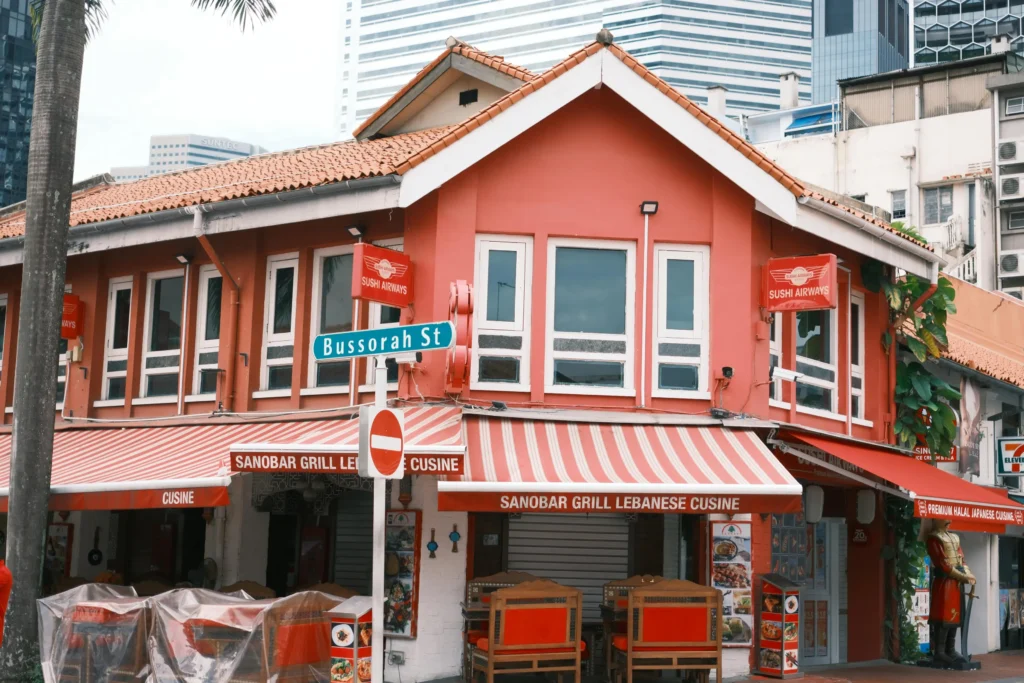 A vibrant two-story, terracotta-orange shophouse stands at the corner of Bussorah Street, featuring distinctive white-framed windows and red-and-white striped awnings. The ground floor houses "Sanobar Grill Lebanese Cuisine" with outdoor seating, while red signs for "Sushi Airways" are visible on the upper level against a backdrop of modern skyscrapers.