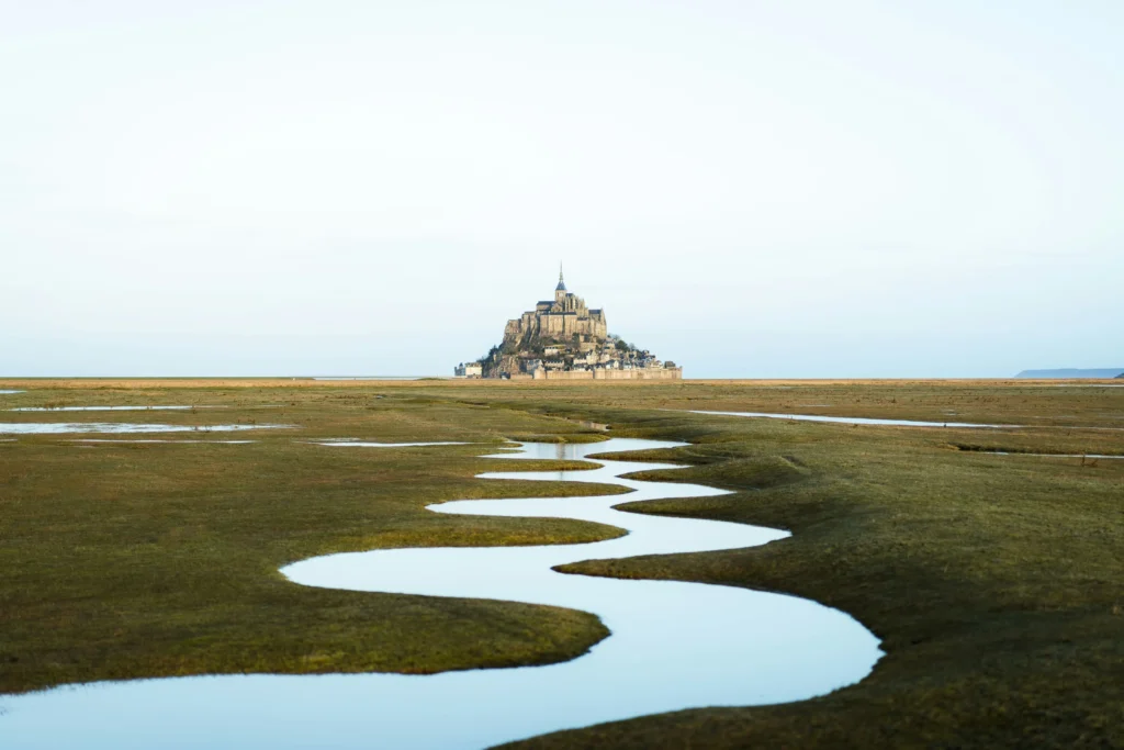In the image, a winding, S-shaped stream of water cuts through a vast, grassy tidal flat in the foreground. In the far distance, the historic island abbey of Mont Saint-Michel stands prominently under a wide, pale sky.