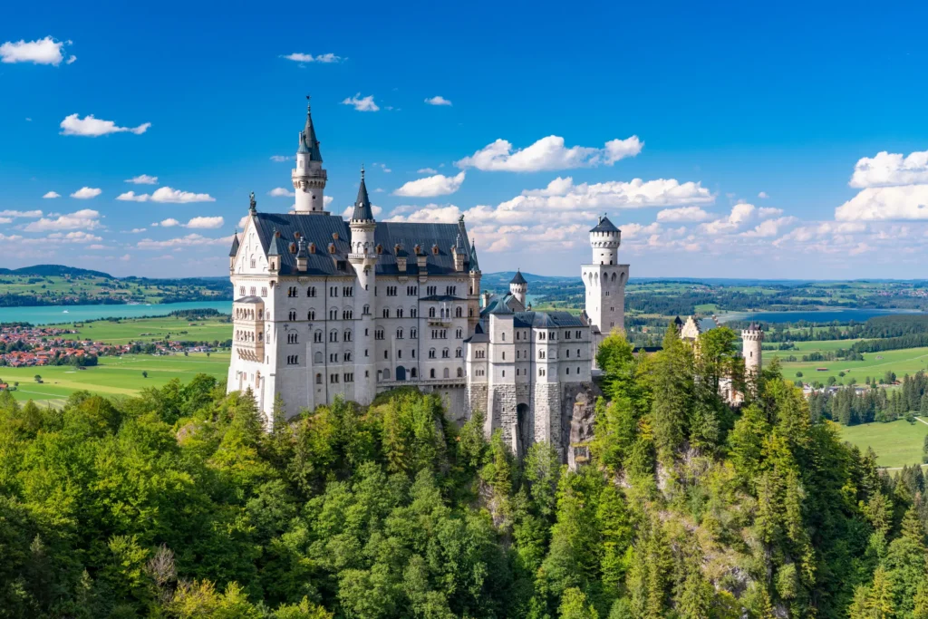 A fairy-tale-style white castle with dark pointed turrets sits atop a lush, tree-covered hill under a bright blue sky with scattered clouds. The background features a sweeping landscape of green fields, scattered houses, and distant lakes.