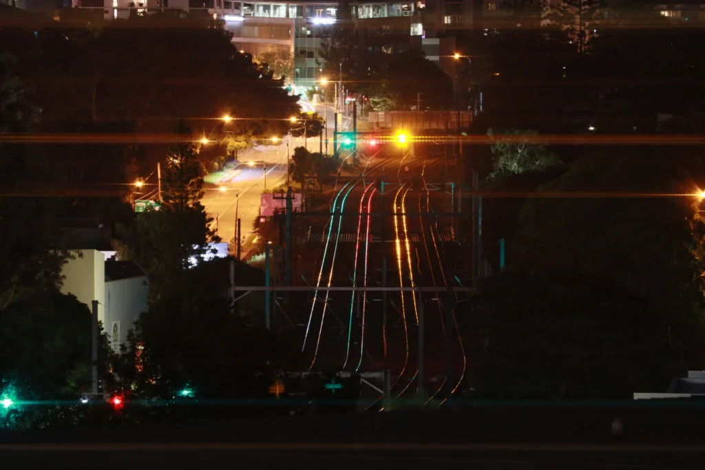 The image captures a high-angle night view of multiple train tracks curving through an urban landscape surrounded by dense trees. Colorful light streaks from city traffic and glowing railway signals illuminate the dark, contrasting sharply with the night.