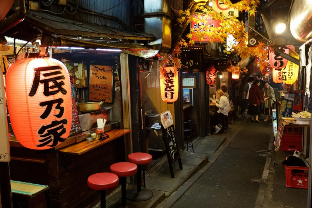 The image captures a narrow, bustling alleyway in Japan lined with traditional food stalls and glowing red paper lanterns. People walk down the alley and dine at small counters under artificial autumn foliage, creating a warm and lively nightlife atmosphere.