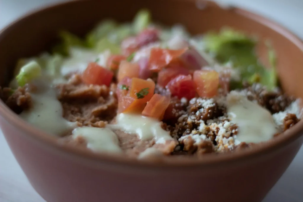 A close-up shot of a Mexican burrito bowl served in a terracotta dish. It is layered with refried beans, ground meat, guacamole, shredded lettuce, melted cheese, and fresh pico de gallo.