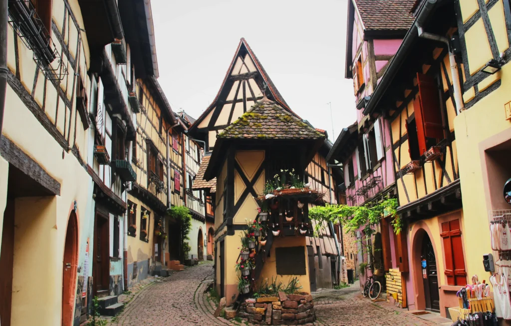 The image shows a narrow, charming cobblestone street lined with colorful, traditional half-timbered houses. In the center, the path forks around a small, quaint building decorated with potted flowers and a steeply pitched roof.