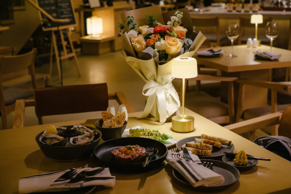 A beautifully arranged dinner table in a dimly lit restaurant features a bouquet of roses, a small cordless lamp, and plates of fresh oysters, steak tartare, and sliced bread. The background shows a warm, inviting dining area with wooden chairs and additional tables under soft lighting.