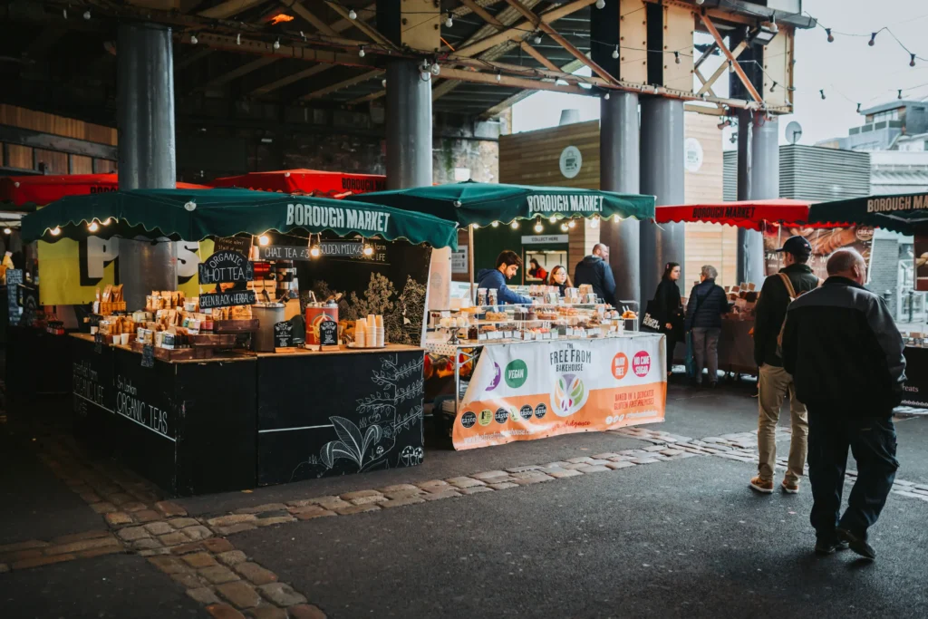 The image displays several food stalls with dark green awnings labeled "Borough Market" operating underneath a large overhead bridge structure. Vendors serve customers from counters filled with organic teas and baked goods while pedestrians walk across the paved ground.