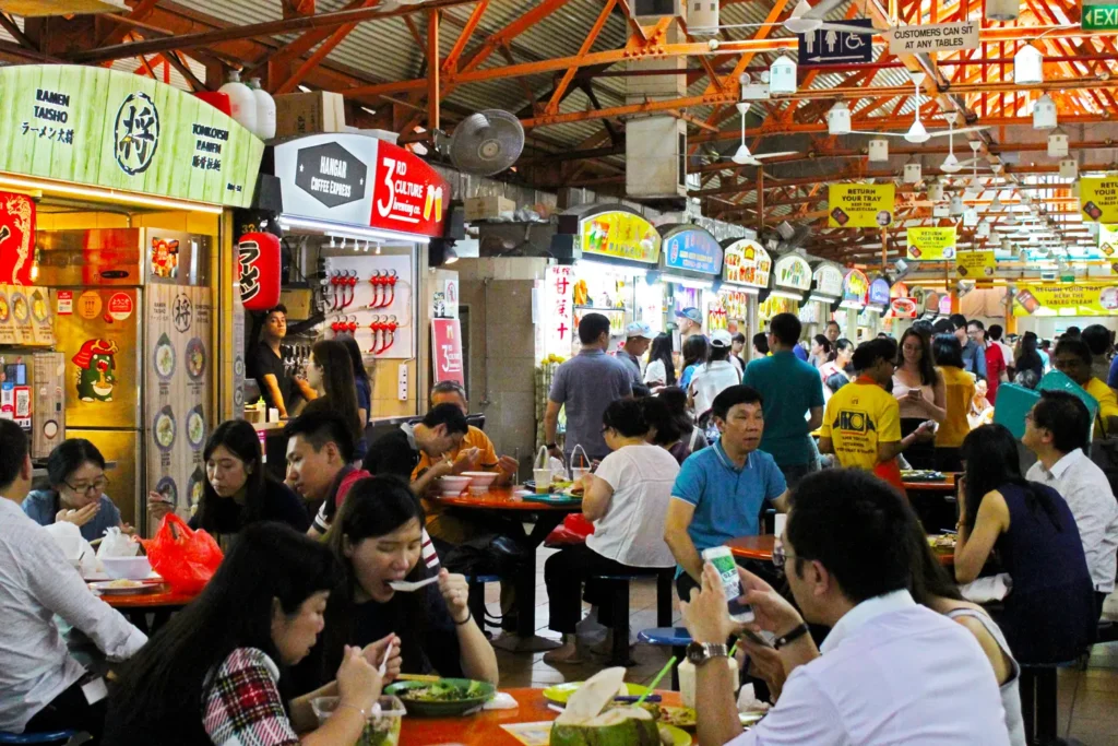 A bustling Singaporean hawker centre is filled with people seated at round tables enjoying meals amidst a row of vibrant food stalls. Overhead, bright yellow signs remind patrons to return their trays beneath an open industrial ceiling structure.